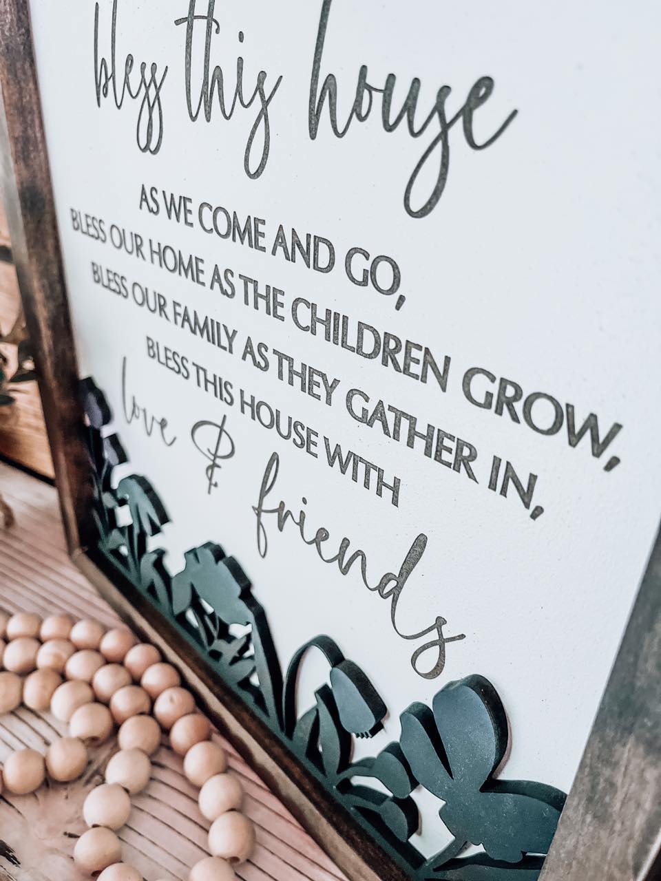 Bless This House farmhouse sign styled on a wooden mantle with white flowers and rustic beads.