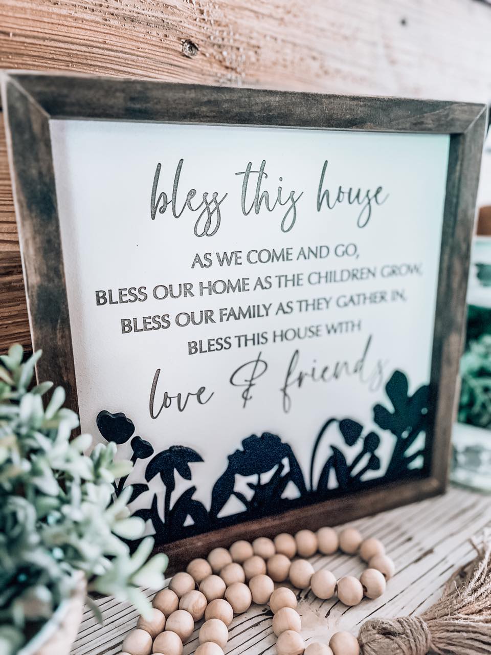 Bless This House farmhouse sign styled on a wooden mantle with white flowers and rustic beads.