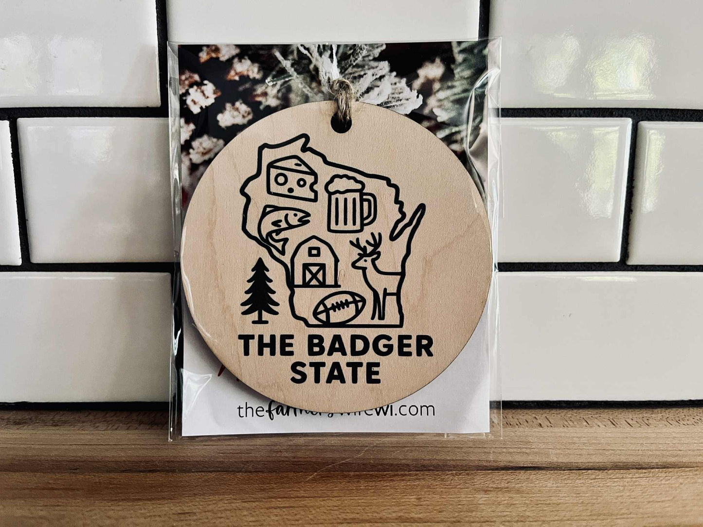Round maple wood ornament with dark green UV-printed Wisconsin design and jute cord, featuring cheese, beer, deer, and farm icons.