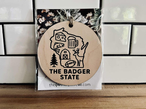 Round maple wood ornament with dark green UV-printed Wisconsin design and jute cord, featuring cheese, beer, deer, and farm icons.