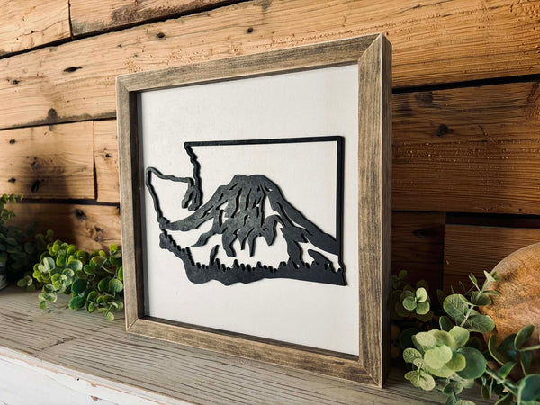 Framed black cutout of a mountain on a wooden wall with plants.