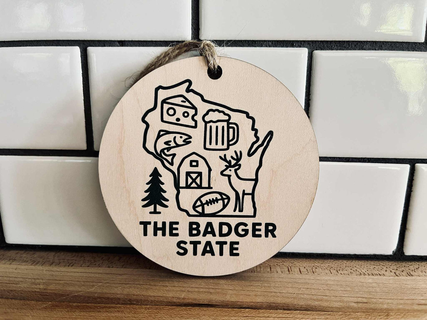 Round maple wood ornament with dark green UV-printed Wisconsin design and jute cord, featuring cheese, beer, deer, and farm icons.