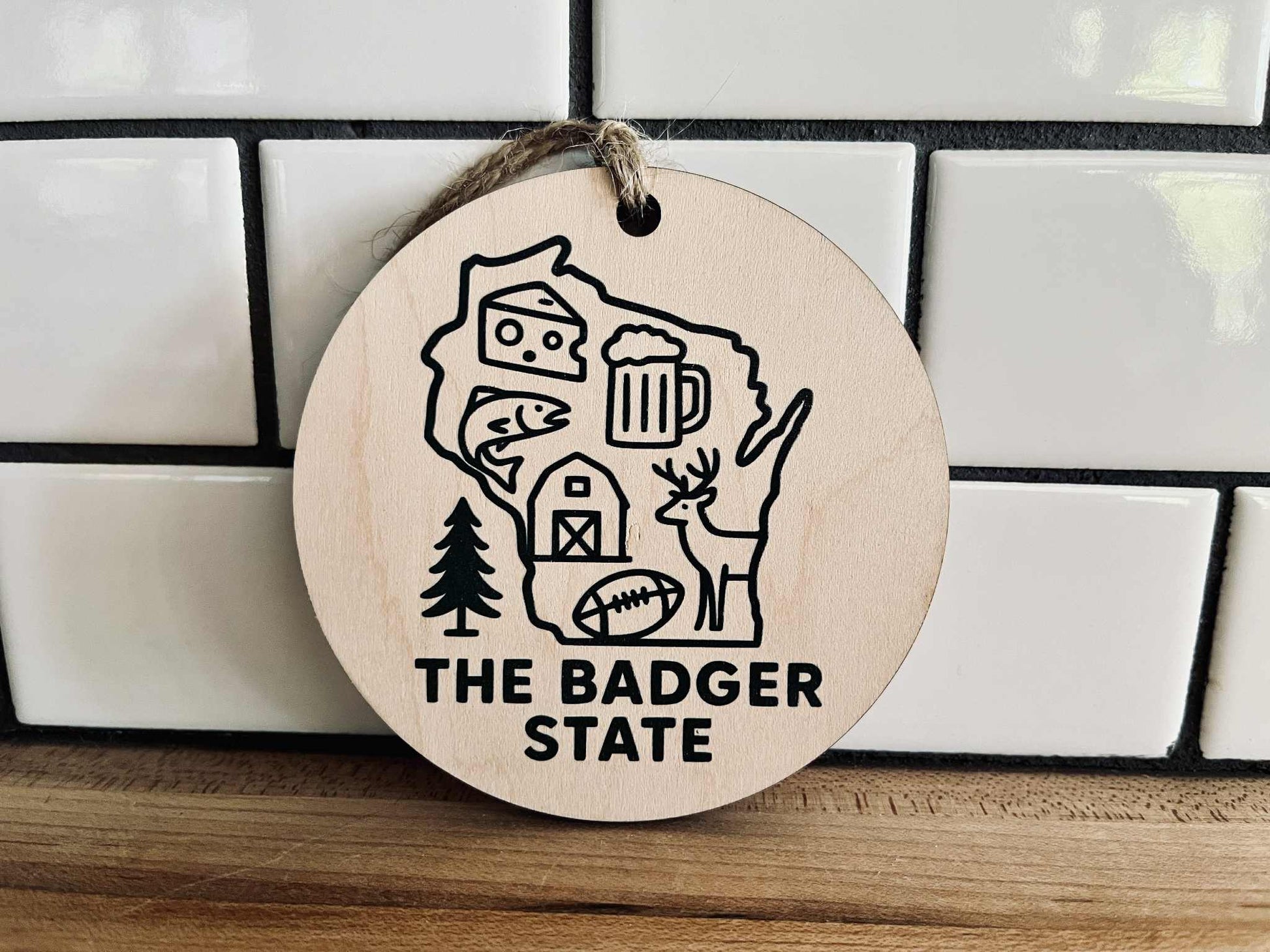 Round maple wood ornament with dark green UV-printed Wisconsin design and jute cord, featuring cheese, beer, deer, and farm icons.