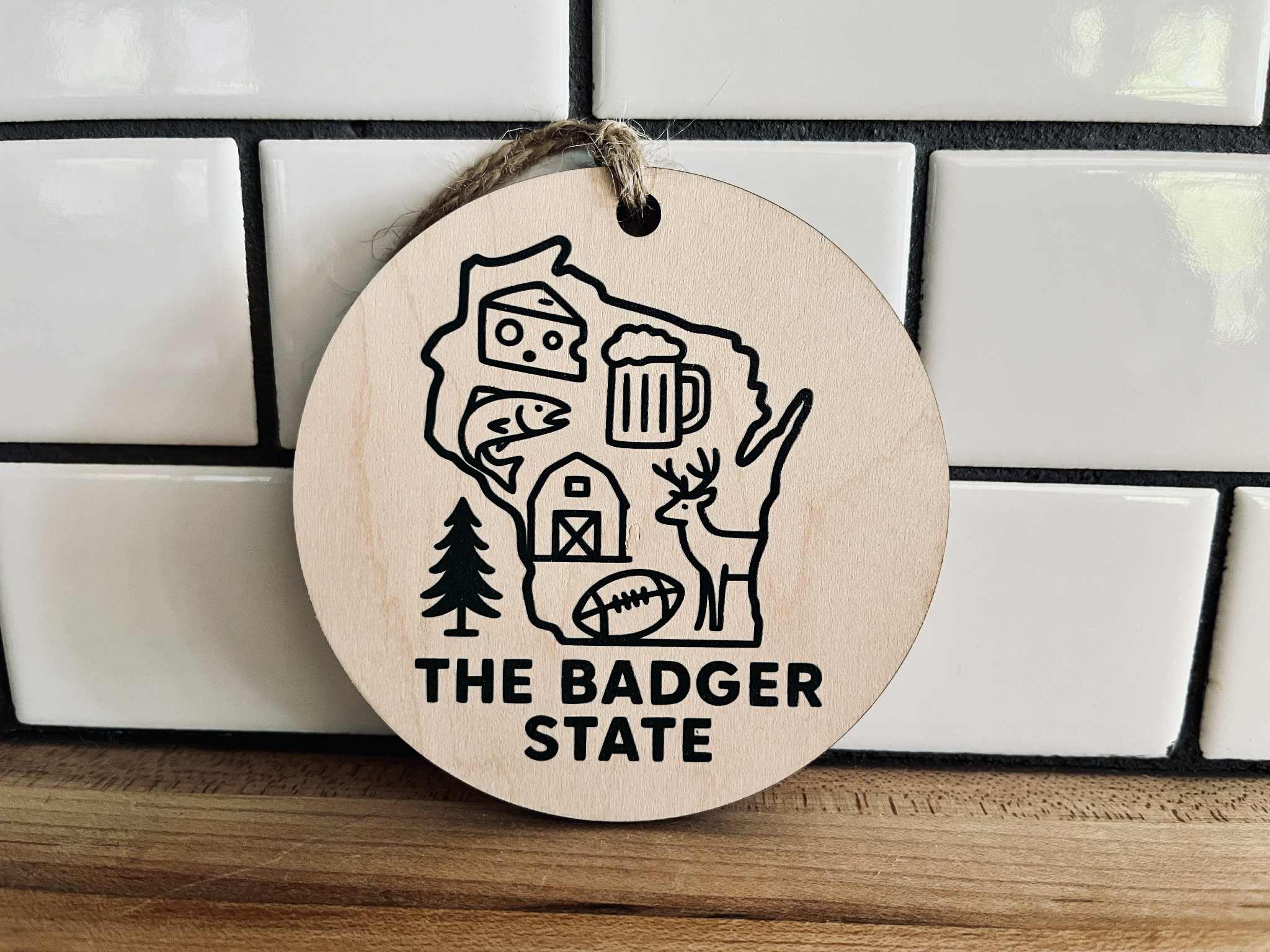Round maple wood ornament with dark green UV-printed Wisconsin design and jute cord, featuring cheese, beer, deer, and farm icons.