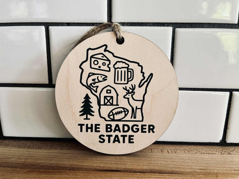 Round maple wood ornament with dark green UV-printed Wisconsin design and jute cord, featuring cheese, beer, deer, and farm icons.